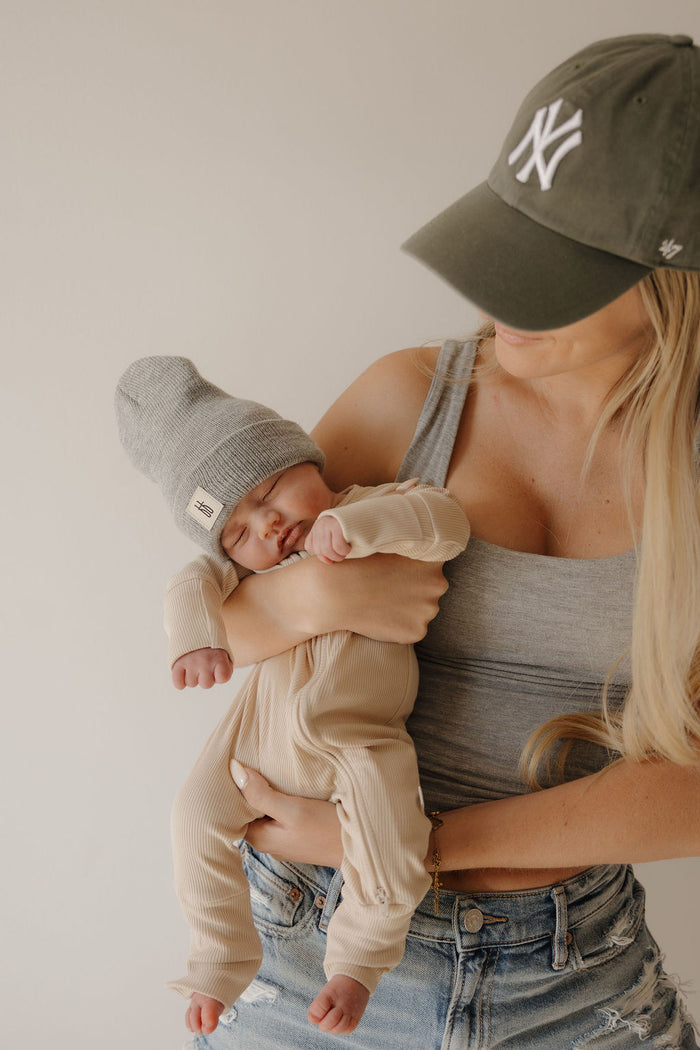 A woman in a gray tank top and NY cap holds a sleeping baby wearing forever french baby's Bamboo Zip Pajamas | Ribbed Cream and a gray knit hat, both softly lit to highlight the pajamas' premium bamboo comfort.