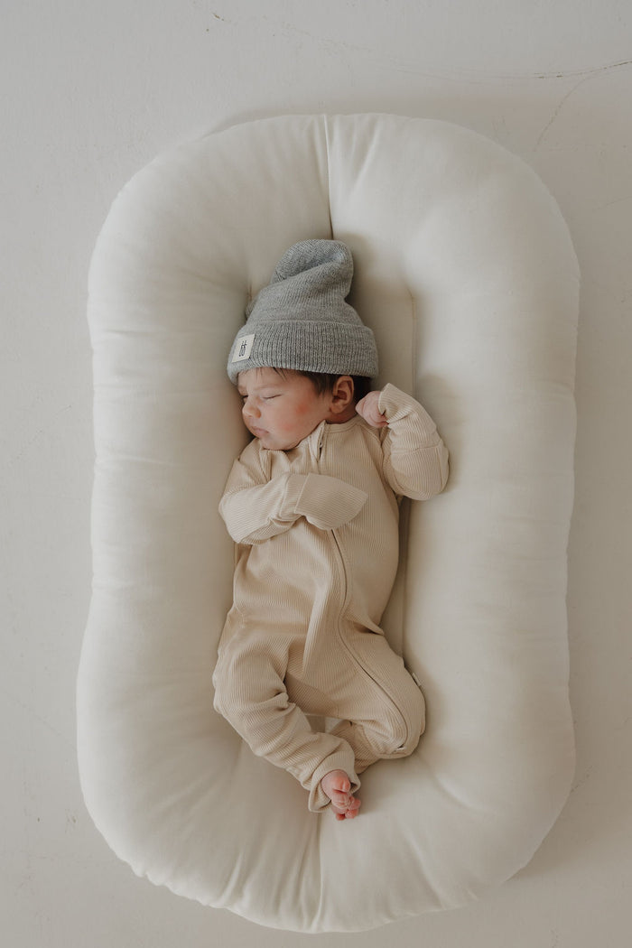 A newborn sleeps on a soft, white, oval cushion, wearing forever french baby’s Bamboo Zip Pajamas in ribbed cream and a gray knit hat with a small label, lying peacefully with hands near the face.