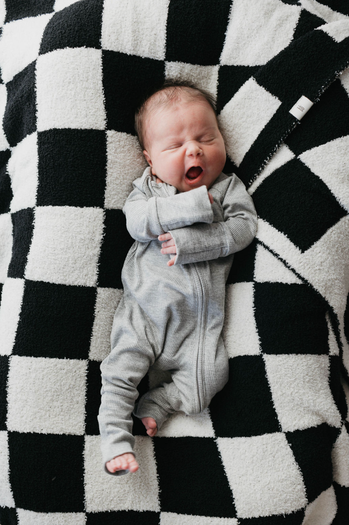 A newborn in Bamboo Zip Pajamas | Ribbed Grey by forever french baby lies on a black and white checkered blanket, exuding cozy charm. The baby appears to be yawning with eyes closed and mouth open.