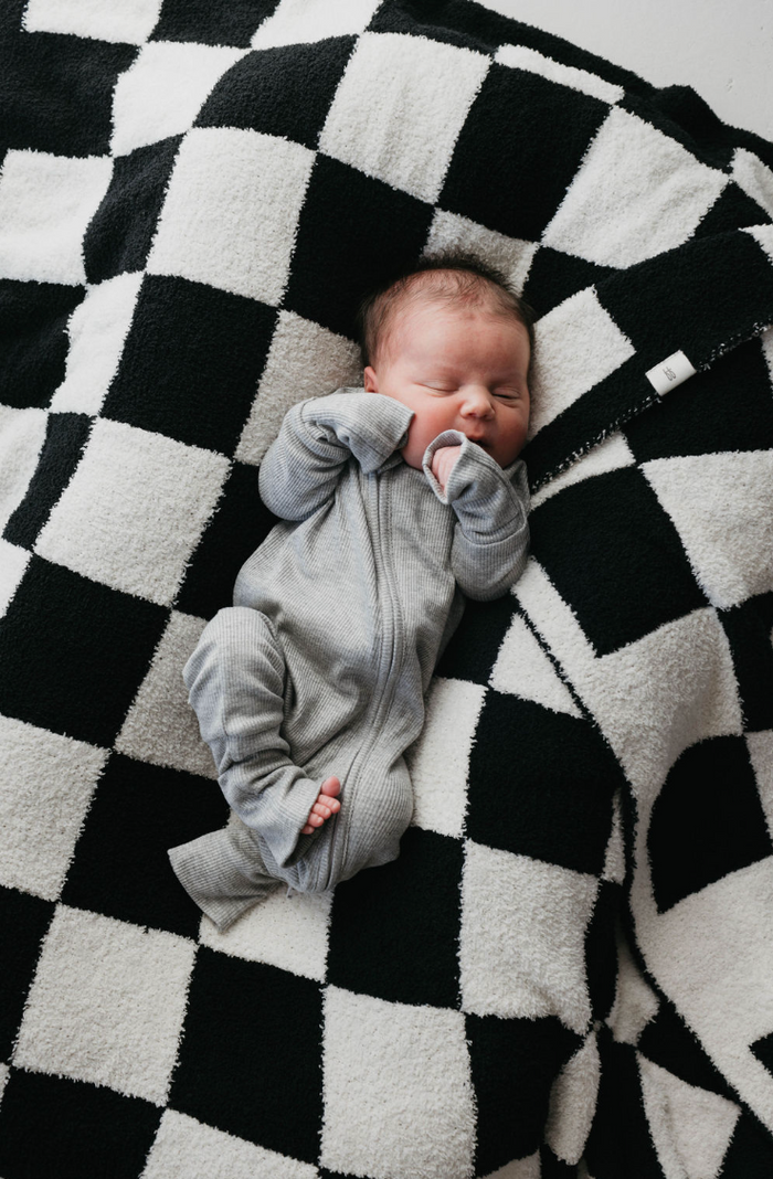 A baby in Bamboo Zip Pajamas (Ribbed Grey) by forever french baby rests peacefully on a black and white checkered blanket, surrounded by timeless style.