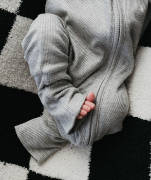 A baby wearing forever french baby's Ribbed Grey Bamboo Zip Pajamas lies on a black and white checkered surface, their tiny toes peeking from the pant leg.