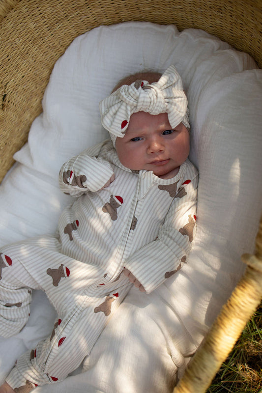 A baby wearing forever french baby's Bamboo Zip Pajamas | Santa Teddy, featuring a soft striped design and a matching headband, lies in a woven basket lined with a white blanket, gazing upward.