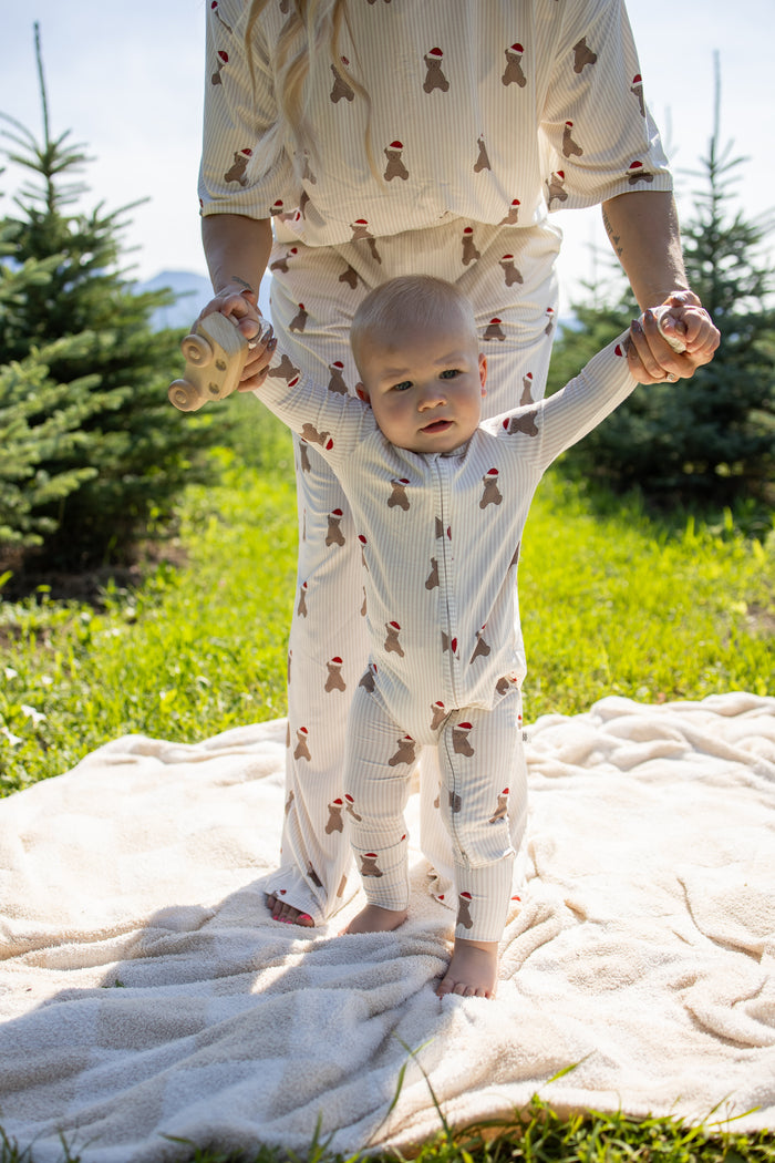 Clad in forever french baby’s Bamboo Zip Pajamas | Santa Teddy, a baby with matching festive prints is held up by an adult, standing on a blanket outdoors among pine trees on a sunny day.