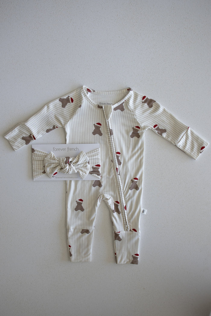 Introducing the Bamboo Zip Pajamas | Santa Teddy by forever french baby: a cozy baby outfit featuring beige and white stripes with charming brown teddy bear and festive red Santa hat print, laid flat. Crafted from hypoallergenic materials gentle for delicate skin. A matching bow headband is packaged to the left of the romper for a coordinated look.