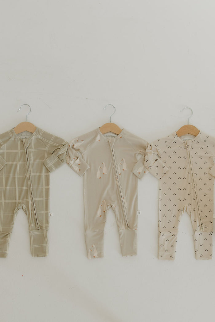 Three Bamboo Zip Pajamas by forever french baby hang on wooden hangers. The left, featuring an olive checkered pattern; the center, beige with animal designs; and the right, cream with small black print. Each onesie includes a central zipper for easy changes against a light background.