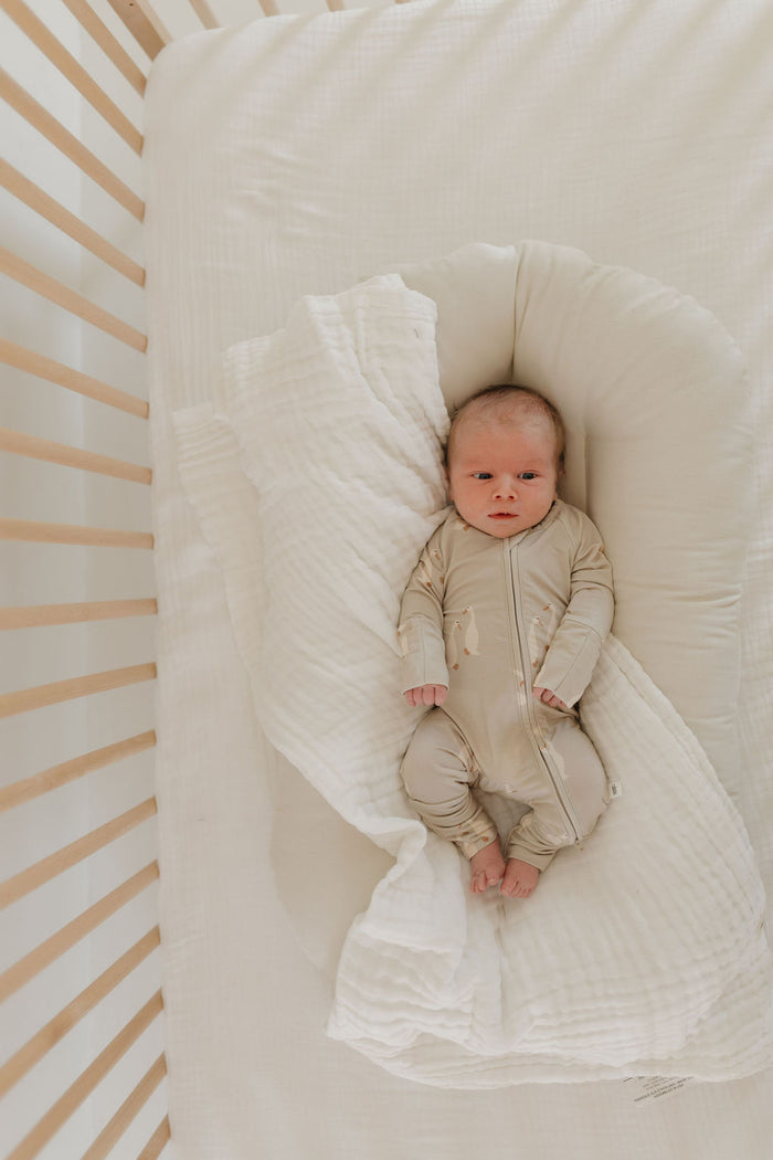 A baby wearing forever french baby's Bamboo Zip Pajamas by Silly Goose lounges awake and curious on a soft white cushion in a crib with pale wooden slats, surrounded by a textured white blanket, creating a serene and cozy setting.