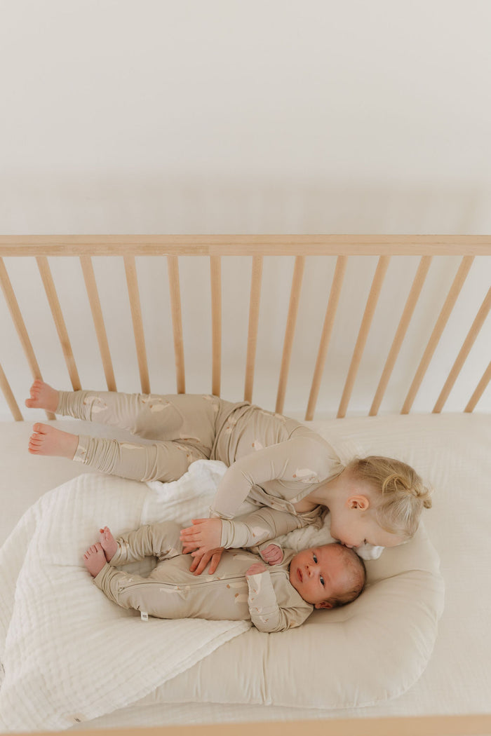 A toddler gently kisses a baby in matching beige Bamboo Zip Pajamas by forever french baby, lying on a soft cushion in a wooden crib with an airy atmosphere that complements their breathable wear perfectly.