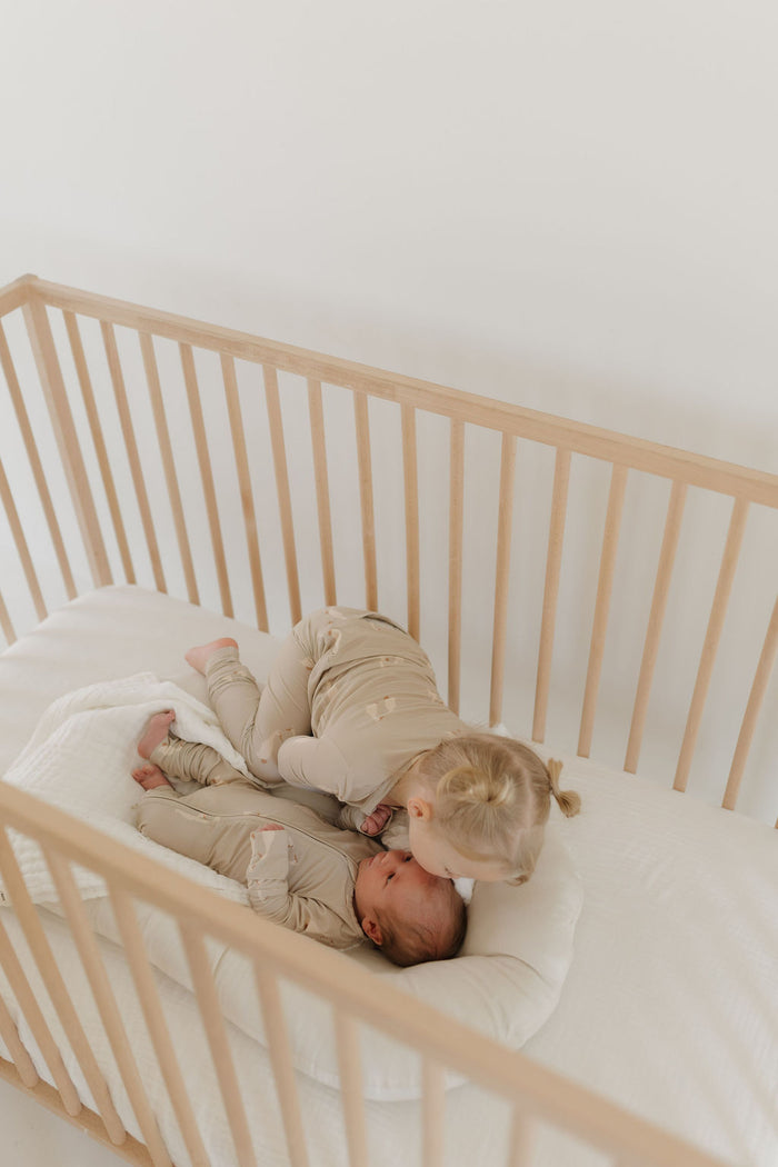 A toddler in Silly Goose Bamboo Zip Pajamas by forever french baby leans over a wooden crib to kiss a baby also wearing soft, hypo-allergenic clothes. The crib holds them on a white mattress, bathed in gentle natural light.