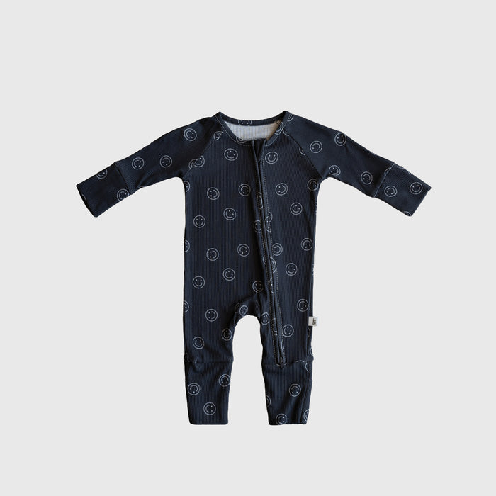 The forever french baby Bamboo Zip Pajamas | Smiley Ribbed feature dark blue ribbed bamboo fabric, a white smiley face print, long sleeves, and a diagonal neck-to-leg zipper, shown on a light gray background.