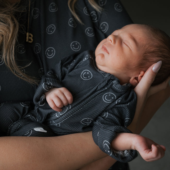 A newborn sleeps peacefully in someone's arms, wearing forever french baby's Bamboo Zip Pajamas | Smiley Ribbed. An adult supports the baby, with part of a necklace visible on their neck.