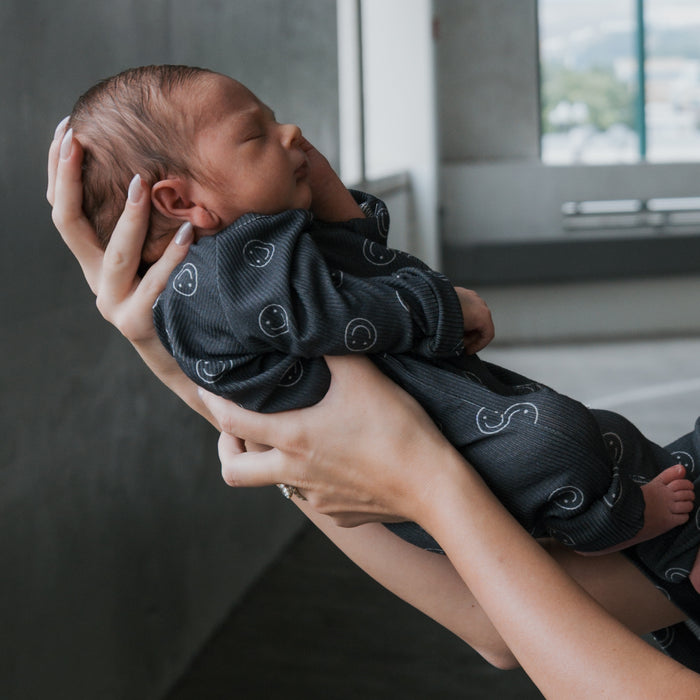 A newborn, gently cradled in adult hands, wears forever french baby's Bamboo Zip Pajamas | Smiley Ribbed, set against a softly blurred indoor background.