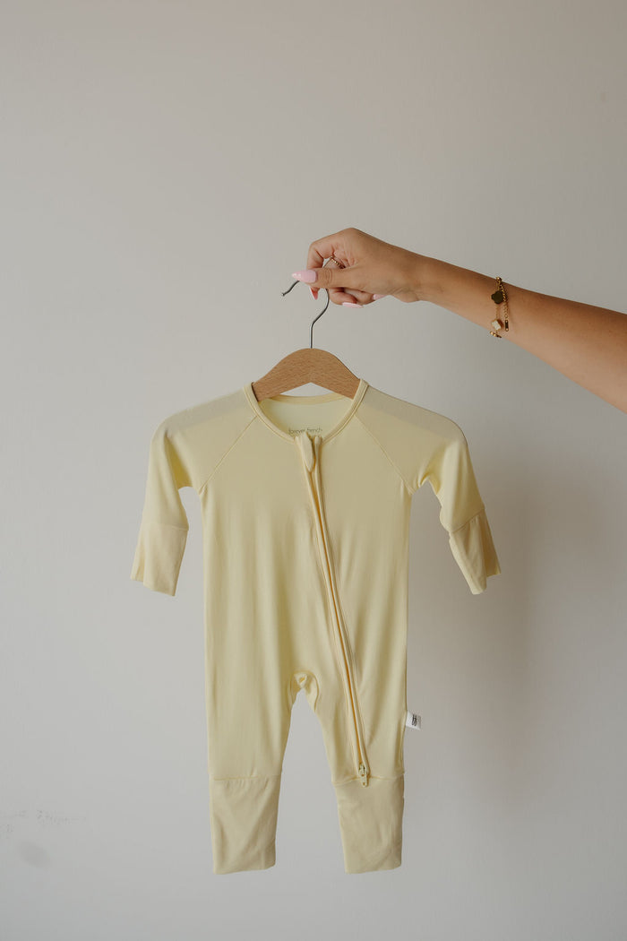A hand holds a wooden hanger displaying the “Bamboo Zip Pajamas | Sunny” by forever french baby, a pale yellow long-sleeve baby onesie with a front zipper, set against a plain light background and highlighting its soft, hypoallergenic fabric that’s ideal for sensitive skin.