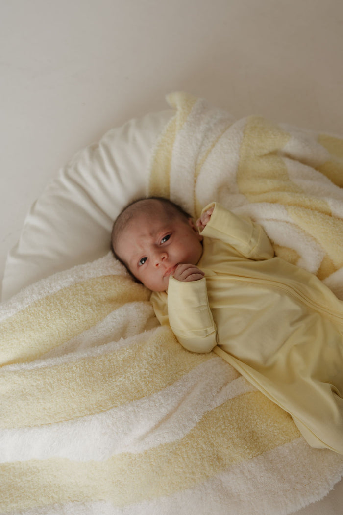 A baby in a yellow outfit lies on a soft, yellow-and-white striped blanket, gazing upward with one hand near their face. The cozy ensemble features the Bamboo Zip Pajamas | Sunny from forever french baby, crafted from breathable bamboo fabric to ensure comfort and gentleness for delicate skin.