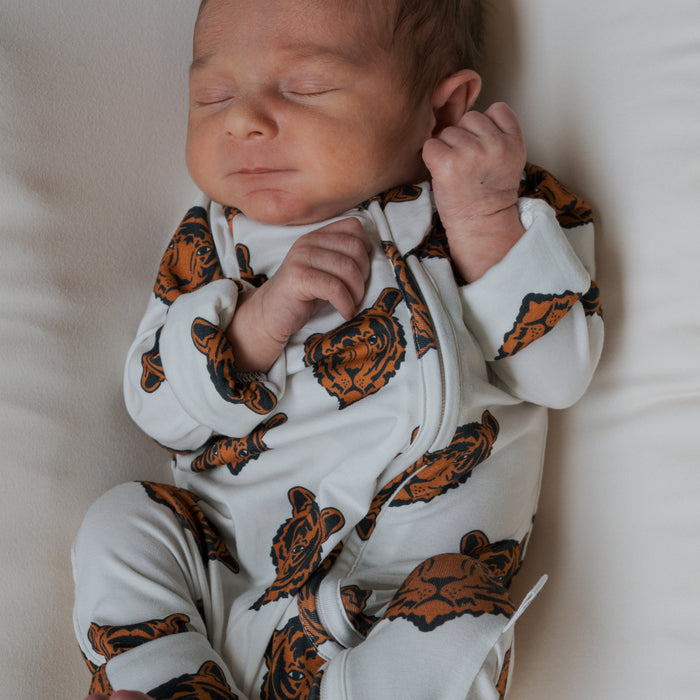 A newborn sleeps peacefully on a white surface, wearing forever french baby’s Bamboo Zip Pajamas in the Tigers print—a soft, breathable onesie decorated with orange and black tiger faces.