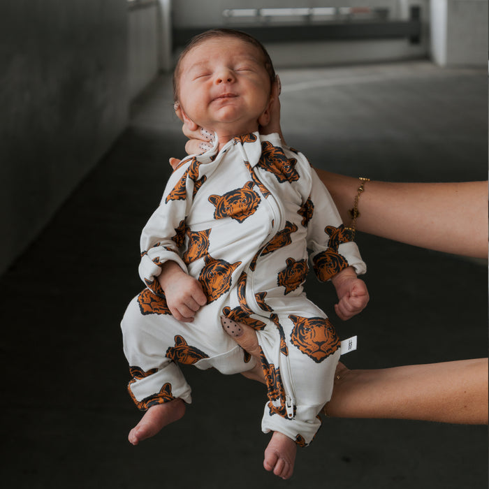 A newborn sleeps peacefully in forever french baby Bamboo Zip Pajamas | Tigers, featuring soft, breathable fabric with tiger faces, gently held by an adult in a dimly lit room.