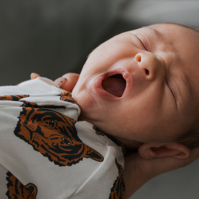 A close-up of a yawning baby in forever french baby's Bamboo Zip Pajamas | Tigers, featuring a tiger face print, being gently held by someone’s hand.