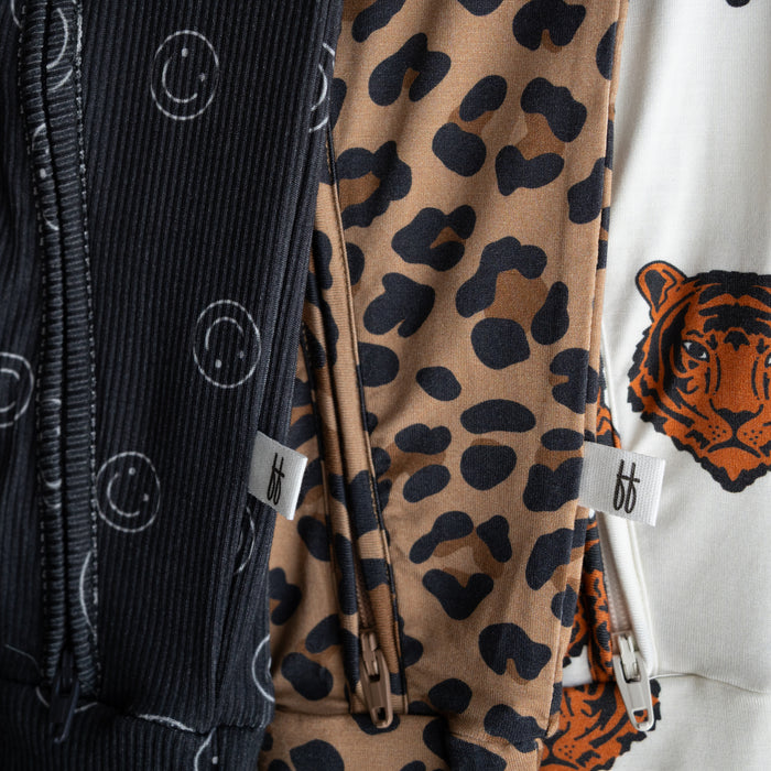 Close-up of three forever french baby Bamboo Zip Pajamas: black with white smiley faces, brown with black leopard spots, and white with a tiger face. Each features soft, breathable fabric and a small white "ff" tag near the zipper.