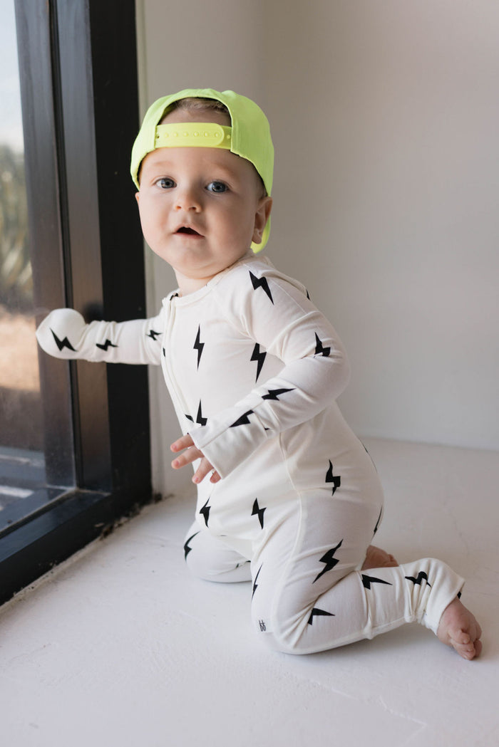 A baby wearing a Bamboo Zip Pajamas in white with black lightning bolt designs from forever french baby and a bright yellow-green baseball cap is kneeling by a window, looking towards the camera. The baby has one hand resting against the window pane, showcasing the unique style of forever french baby.
