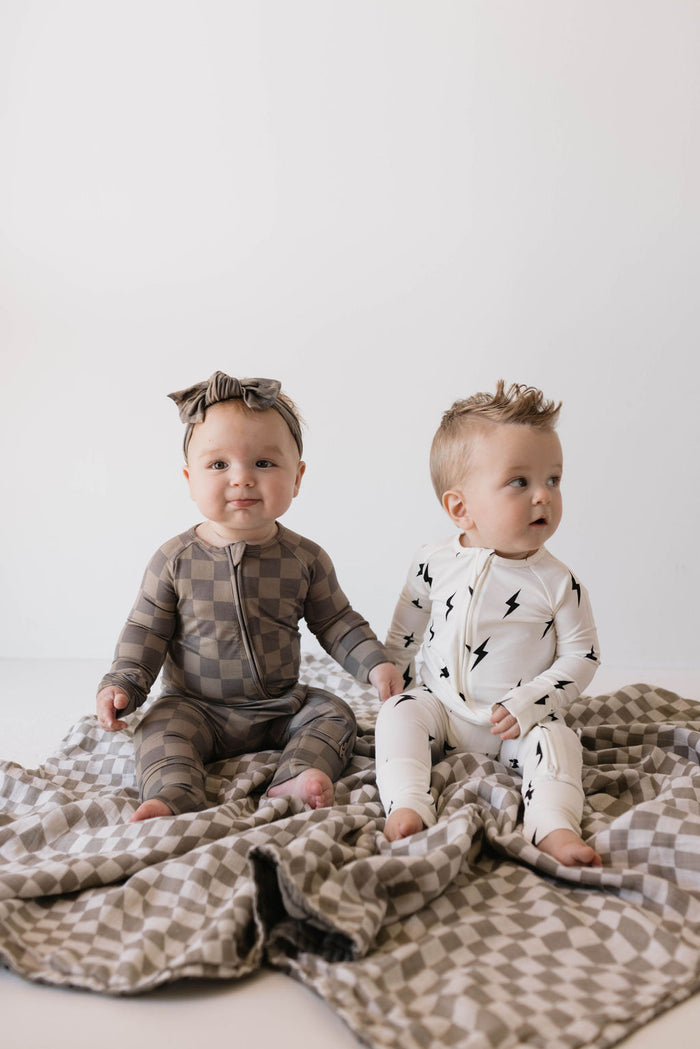 Two babies sit on a checkered blanket. The baby on the left, dressed in a gray and brown checkered outfit with a matching headband, looks adorably cozy. The baby on the right sports Bamboo Zip Pajamas featuring a trendy white and black lightning bolt design from forever french baby. Both babies have light hair and are barefoot.