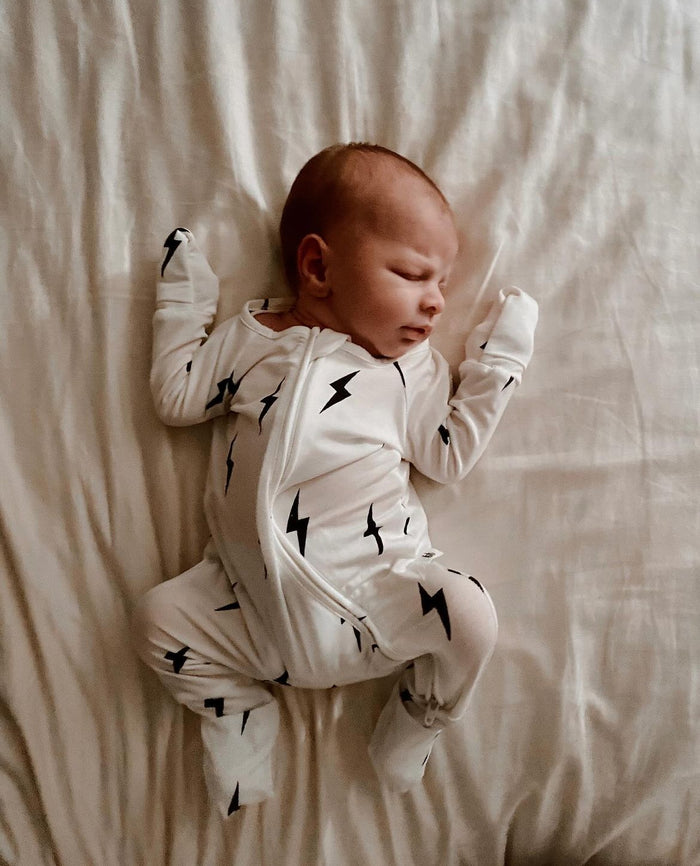 A baby wearing Forever French Baby's Bamboo Zip Pajamas in a chic white and black lightning bolt design lies asleep on a white bed. Positioned on their back with arms slightly raised and legs bent, the baby is nestled on lightly wrinkled bedding, creating a cozy and peaceful atmosphere that reflects Forever French Baby's signature style.
