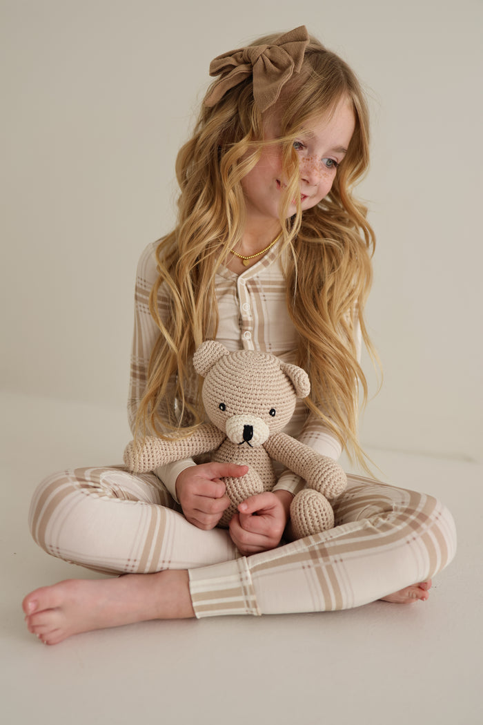 A young girl with long blonde hair and a large brown bow sits cross-legged in plaid pajamas, smiling as she holds the Benson the Bear knit doll by forever french baby in her lap against a plain light background.