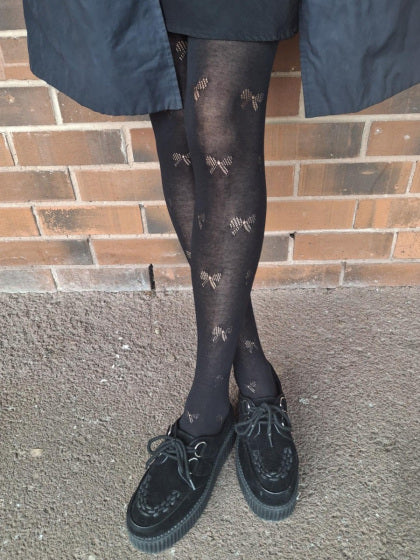 Person wearing black tights with bow patterns and black shoes in front of a brick wall.