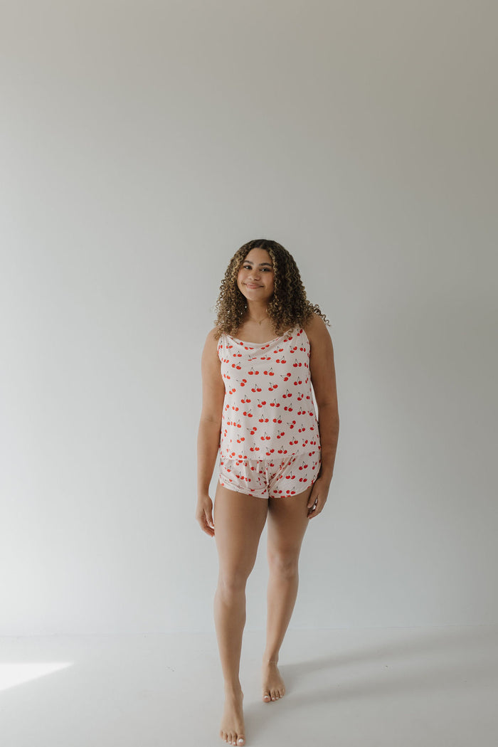 A curly-haired person stands barefoot on a plain, light floor wearing the Cami Women's Bamboo Set by forever french baby. The breathable pajama is hypo-allergenic and features red heart patterns against a minimalist light gray background in the Cherry Orchard design.