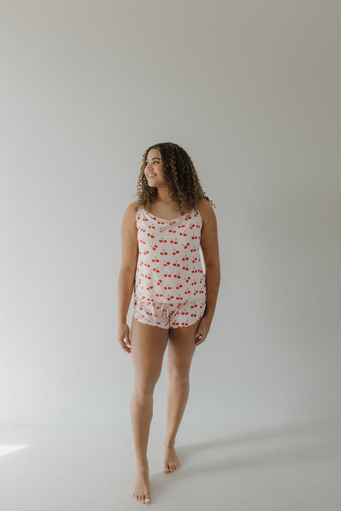 Against a plain backdrop, a person stands barefoot in loose, curly hair wearing the Cami Women's Bamboo Set | Cherry Orchard by forever french baby. This matching sleeveless top and shorts set features a red lips print and is crafted from breathable bamboo fabric under soft diffused lighting.
