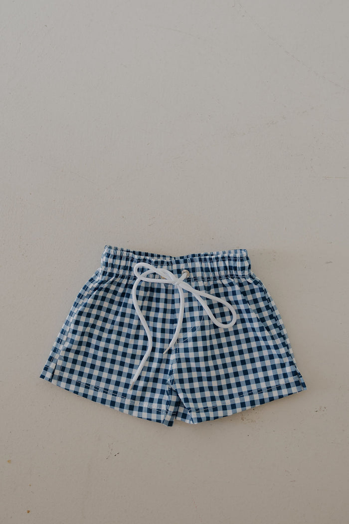 The forever french baby Child Board Short in blue gingham, featuring an elastic waistband and white drawstring, is perfect for summer and shown on a light, textured surface.