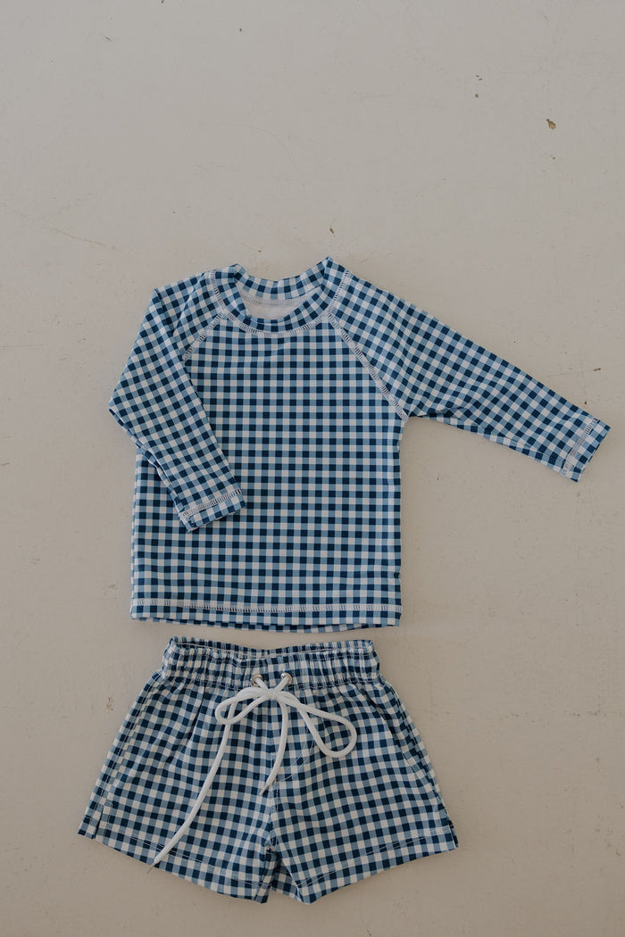 The forever french baby Child Board Short in Blue Gingham features a blue and white check pattern with a long-sleeve top and drawstring shorts, displayed flat on a light background.