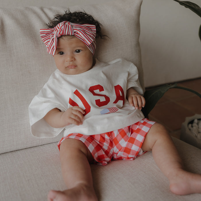 A baby sits on a light couch in a white cotton shirt with "USA" in red, wearing forever french baby's Child Boxer Shorts in Scarlet Gingham and a matching striped bow headband, looking slightly to the side.