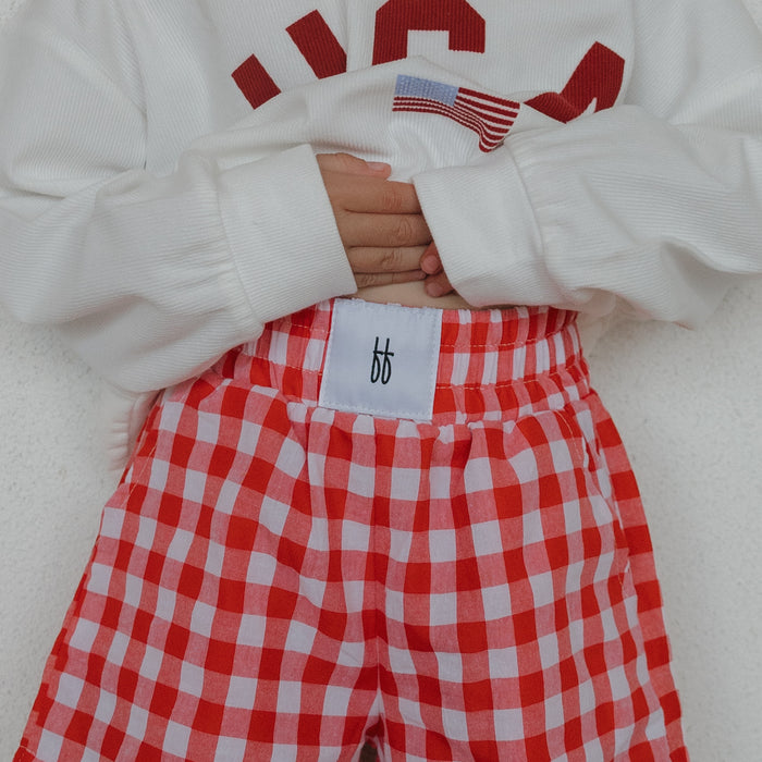 A child wears forever french baby’s Child Boxer Shorts in Scarlet Gingham, paired with a white 100% cotton sweatshirt featuring red lettering, standing against a light background with hands on their stomach.