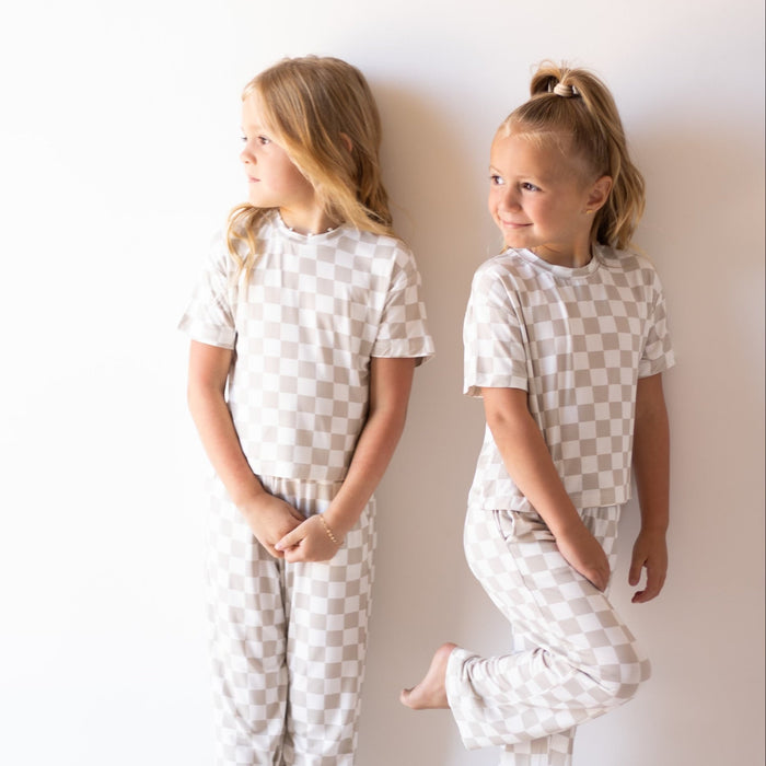Two young girls wear the forever french baby Child Boxy Tee Lounge Set | Checkerboard, standing against a white wall—one barefoot with folded hands looking left, the other balancing on one foot, smiling to the right.