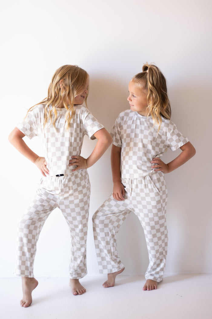 Two young girls with long blonde hair stand barefoot by a white wall, smiling at each other in matching forever french baby Child Boxy Tee Lounge Set | Checkerboard hypo-allergenic pajamas, hands on hips.