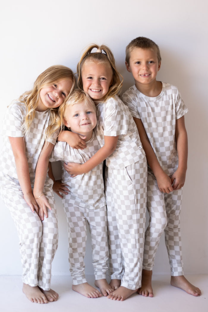 Four young children, all smiling and barefoot, stand together against a white wall wearing forever french baby Bamboo Two Piece Lounge Sets in Checkerboard. One girl hugs another, while the rest stand close and appear happy and relaxed.