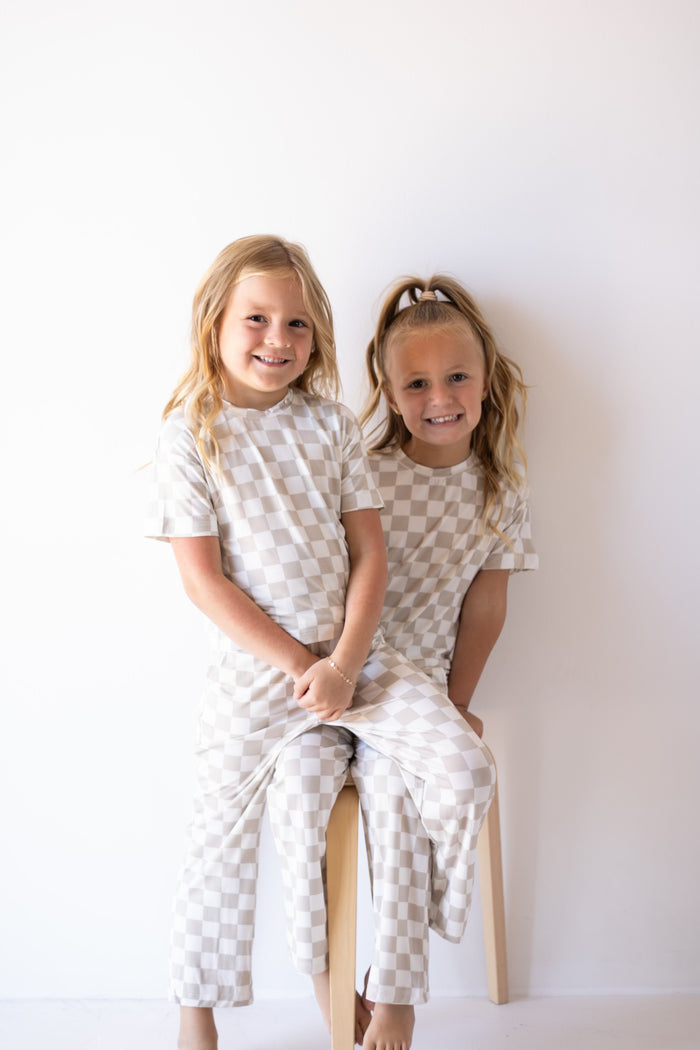 Two young girls with blonde hair, wearing matching forever french baby Child Boxy Tee Lounge Set | Checkerboard, sit smiling together on a light wooden stool against a plain white wall.