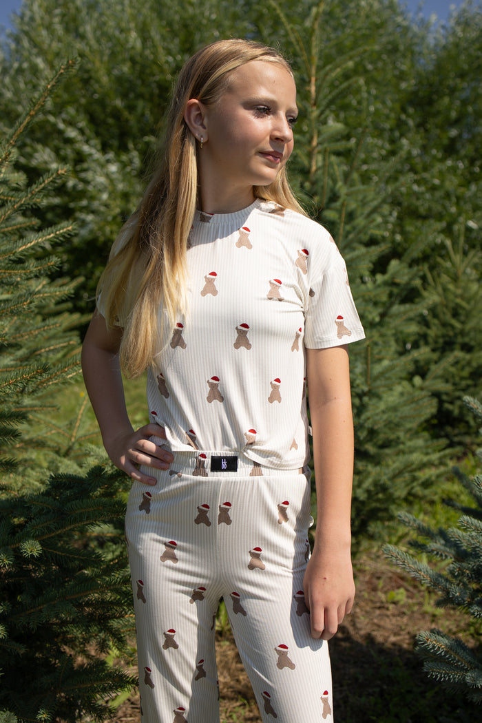 A girl with long blonde hair stands outdoors among pine trees, looking to the side with one hand on her hip. She is wearing the forever french baby Child Boxy Tee Lounge Set in the Santa Teddy print, a light-colored bamboo pajama set featuring small red and brown patterns.