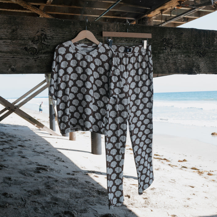 The forever french baby Child Boxy Tee Lounge Set in the Seashells print hangs on wooden hangers under a sunny beach pier, with ocean and sand in the background.