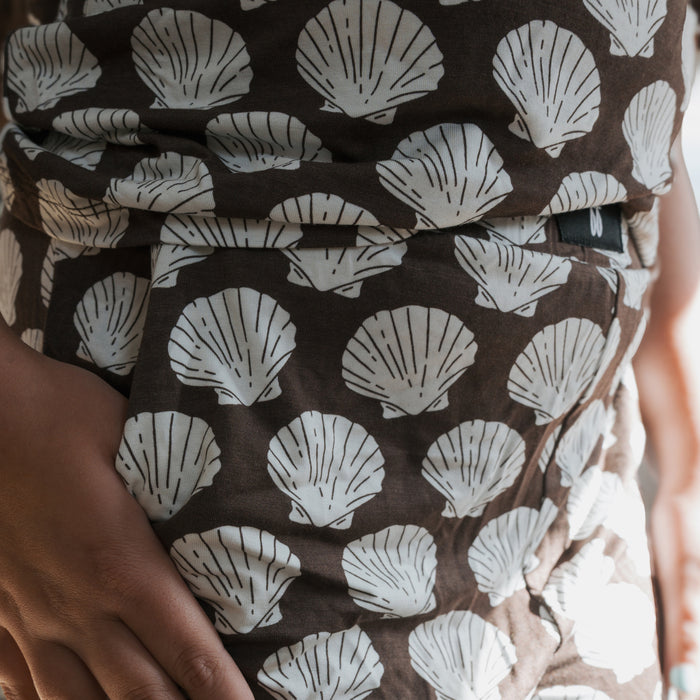 A close-up of a hand resting on a hip wearing the forever french baby Child Boxy Tee Lounge Set in brown, featuring breathable fabric with white seashell patterns highlighted by the light.