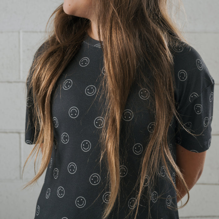 A person with long brown hair wears the forever french baby Child Boxy Tee Lounge Set in Smiley Ribbed, a comfy black bamboo T-shirt with white smiley faces, standing before a light gray brick wall, their face turned away.