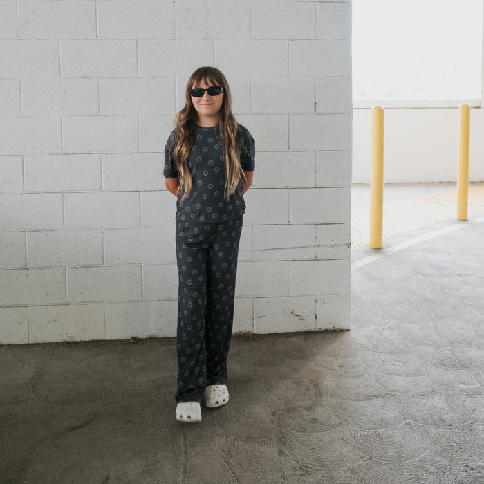 A young person in the forever french baby Child Boxy Tee Lounge Set | Smiley Ribbed stands in front of a white brick wall in a parking garage, sporting long hair, black sunglasses, and white clogs with hands behind their back.