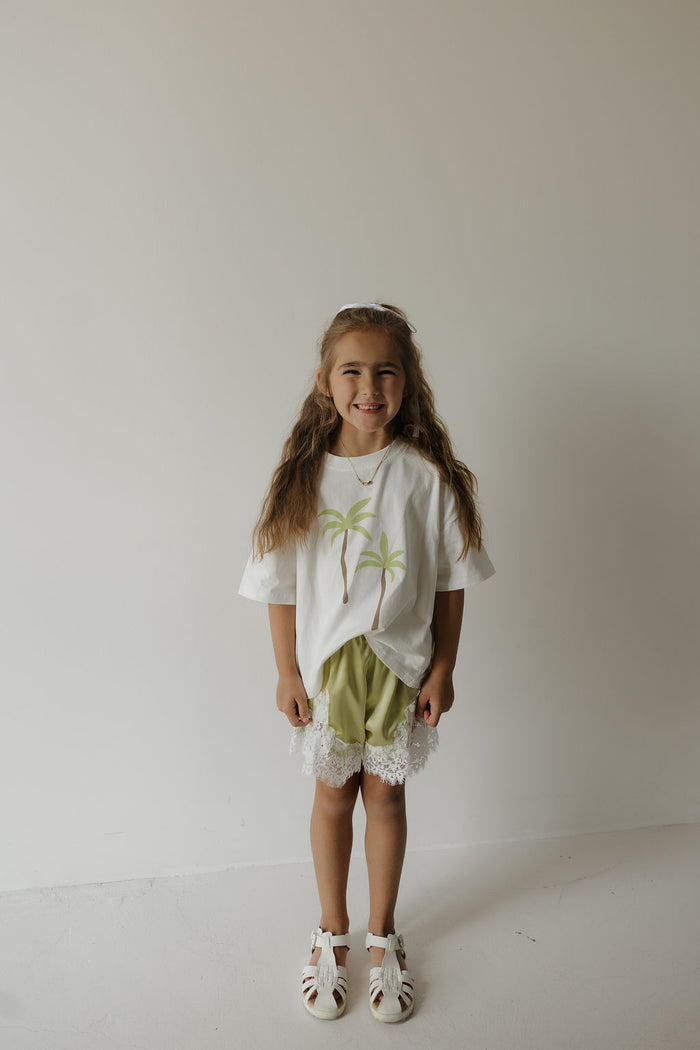 A young girl with long hair stands smiling against a plain white wall, wearing the forever french baby Child Cotton Tee | Palm—crafted from 100% cotton and adorned with palm trees—paired with light green lace-trim shorts and white sandals, making it a perfect choice for coordinated family outfits on sunny days.