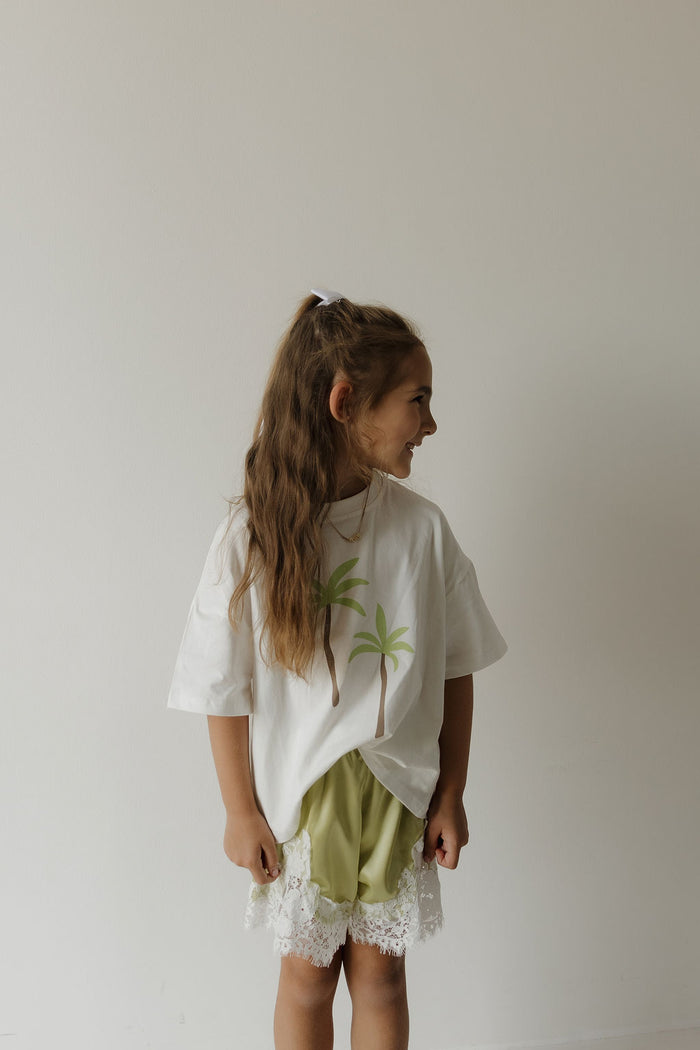 A young girl with long brown hair in a ponytail wears the forever french baby Child Cotton Tee | Palm, featuring palm trees, paired with green lace-trimmed shorts. She stands smiling and looking to the side against a plain white wall.