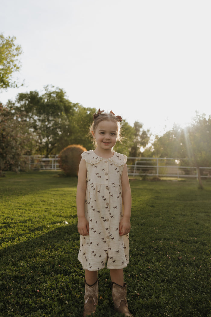 A young girl stands on green grass in a sunlit yard, wearing the forever french baby Child Muslin Dress | Wild Horses. Trees and a fence appear in the background, with sunlight shining from behind her.