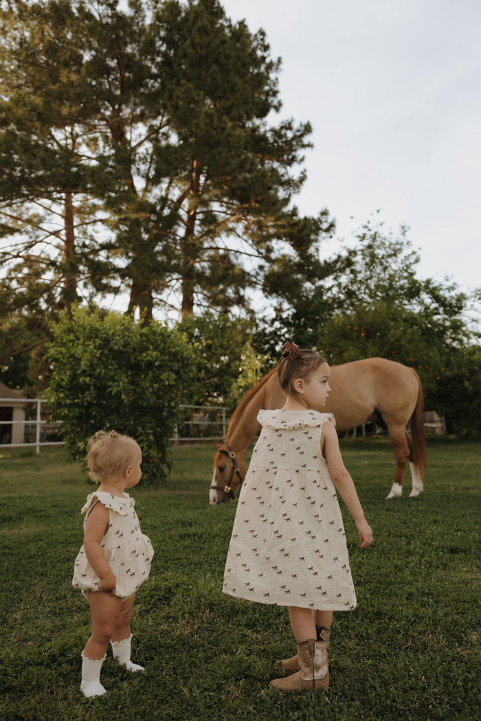 Two young children in matching outfits stand on green grass, watching a tan horse graze. One wears the forever french baby Child Muslin Dress | Wild Horses, blending perfectly with the peaceful outdoor scene.