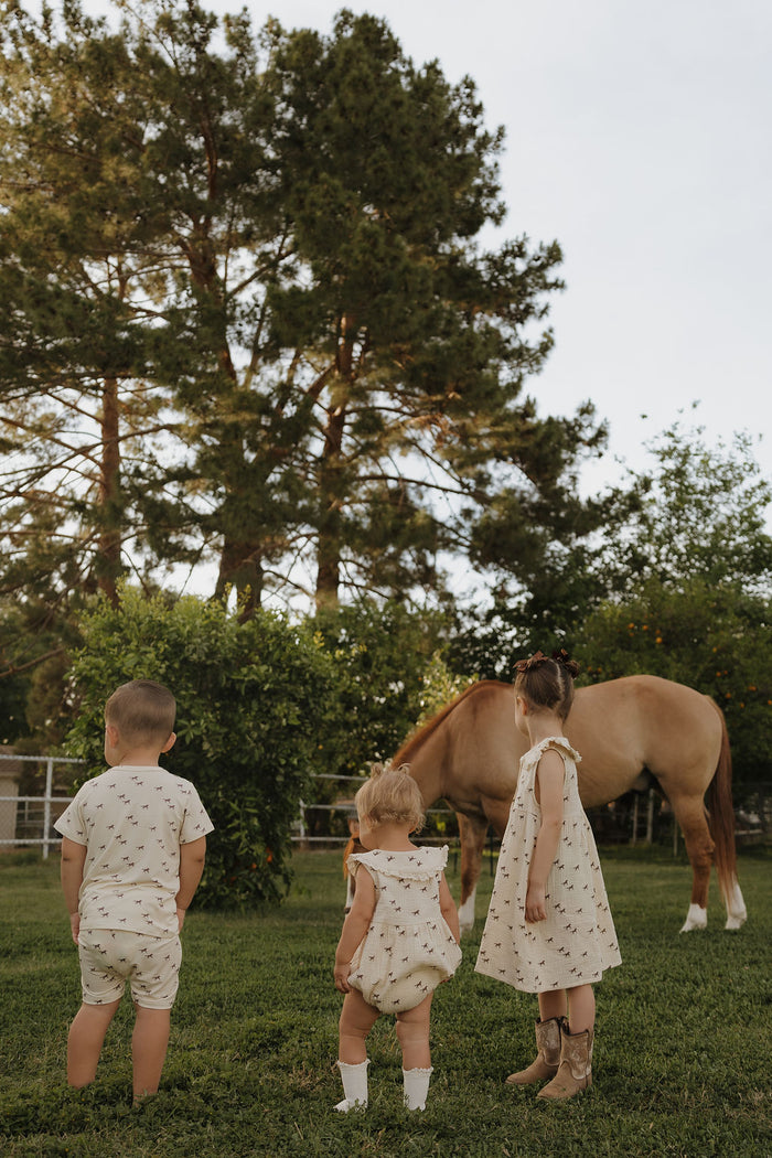 Three children in light, patterned outfits—one wearing the forever french baby Child Muslin Dress | Wild Horses—stand on grass facing a brown horse grazing under tall green trees on a sunny day.