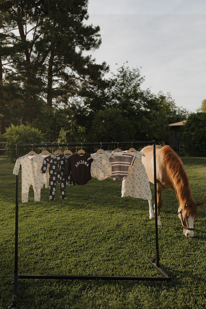 A forever french baby Child Muslin Dress | Wild Horses hangs on a rack with other baby clothes outdoors on grass, as a brown and white horse grazes nearby; trees and bushes are seen under a clear sky.