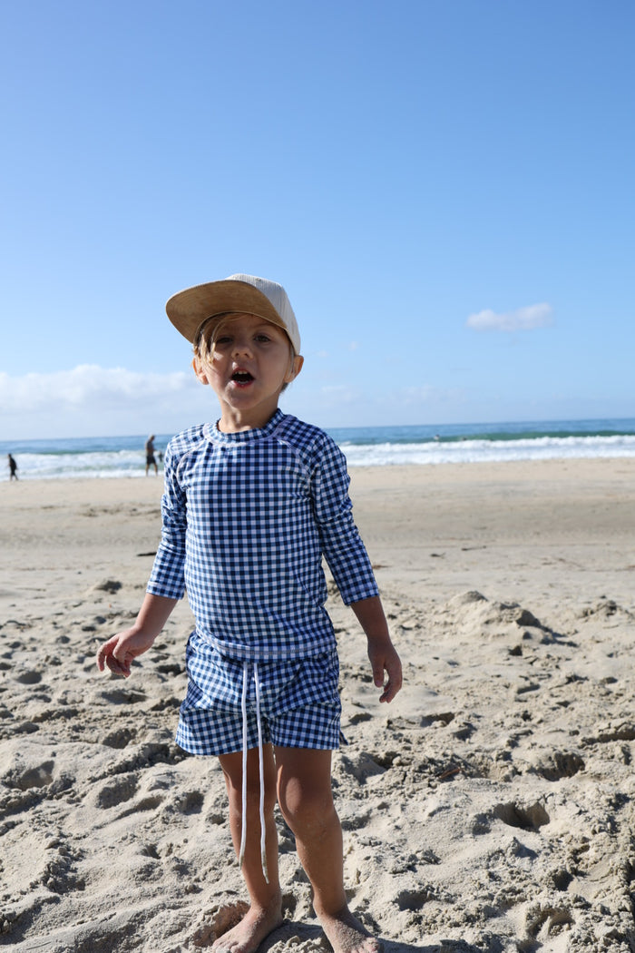 A young child in the forever french baby Child Rash Guard | Blue Gingham stands barefoot on a sandy beach, with the ocean and clear sky behind.