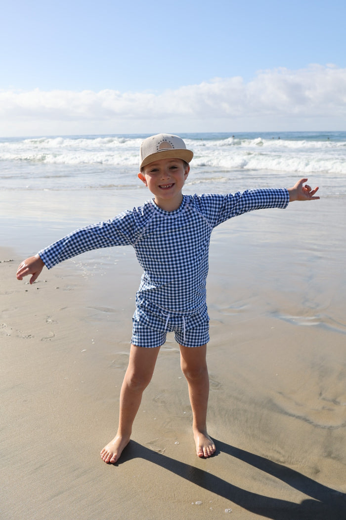 A young child wearing the forever french baby Child Rash Guard | Blue Gingham stands barefoot on the beach with arms outstretched, smiling at the ocean waves in the background.