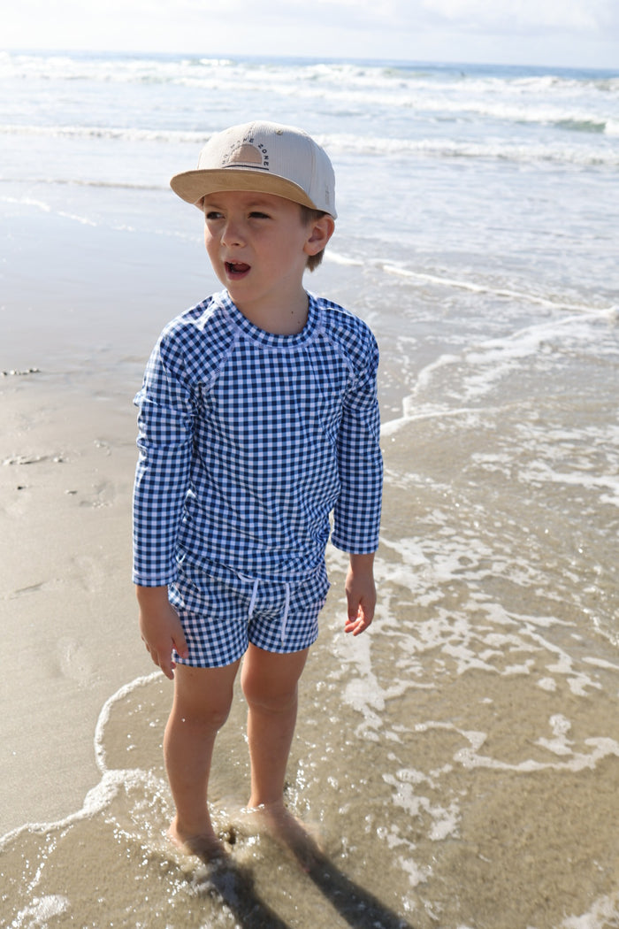 A young boy in the forever french baby Child Rash Guard | Blue Gingham and a beige cap stands barefoot at the ocean’s edge, gentle waves washing over his feet as he enjoys a sunny beach day in perfect summer style.